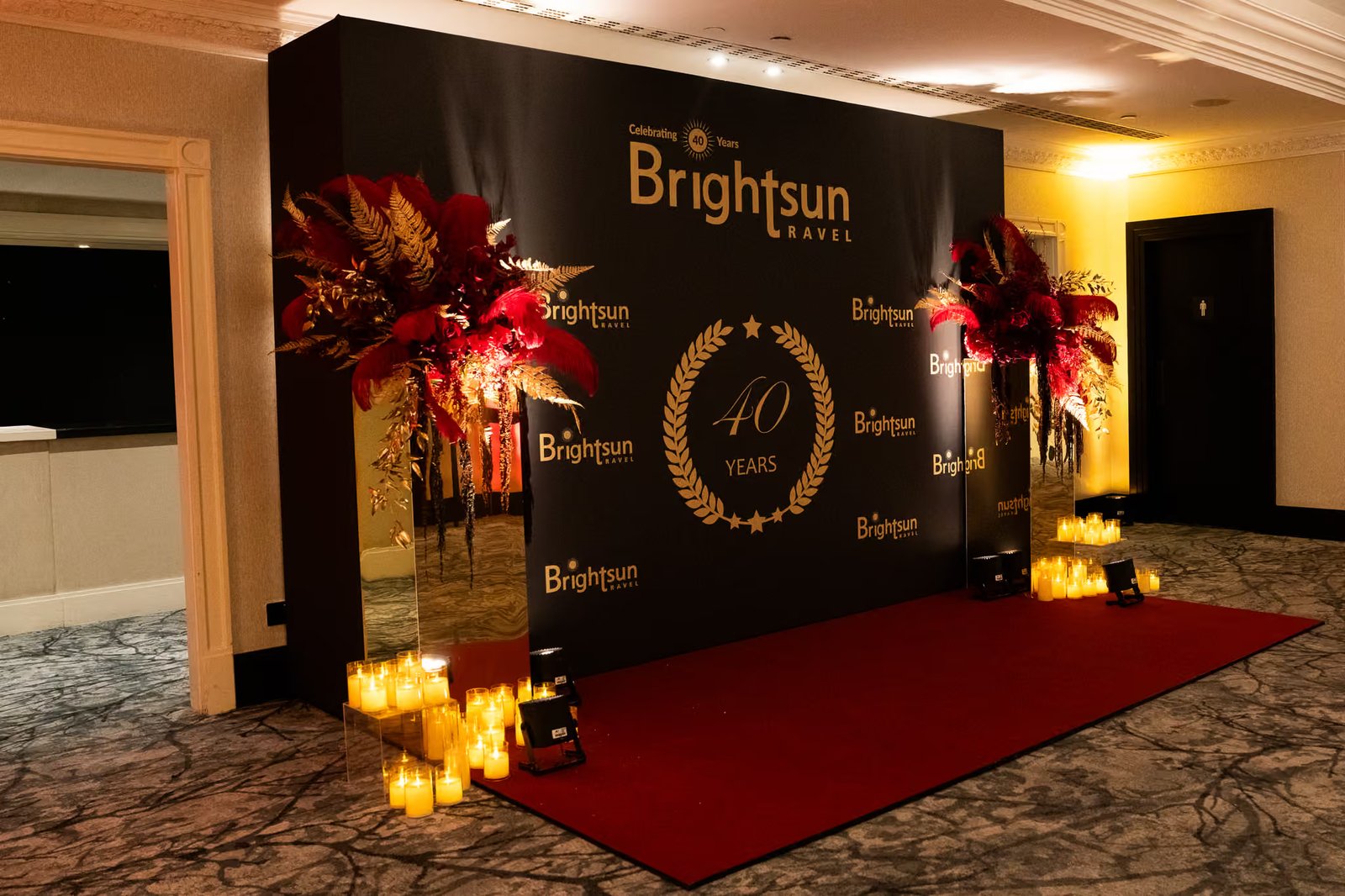 Black and gold Brightsun Travel 40 Years media wall with a red carpet and red floral arrangements