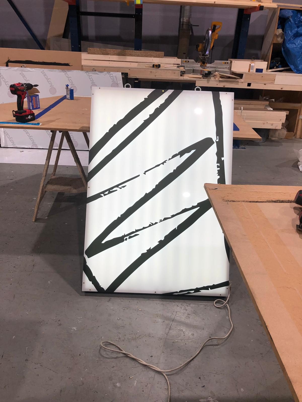 A tall lightbox with bold black brushstroke artwork, glowing in the workshop