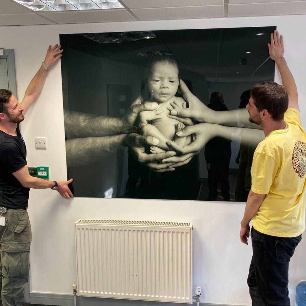 Two people holding up a huge black and white acrylic print of a newborn baby cradled in several pairs of hands
