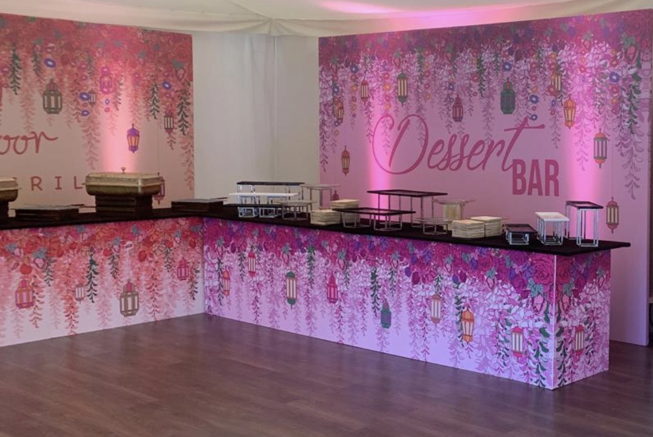 Pink floral printed dessert bar and food counters with matching backdrops covered in cascading flowers and lanterns in an event marquee