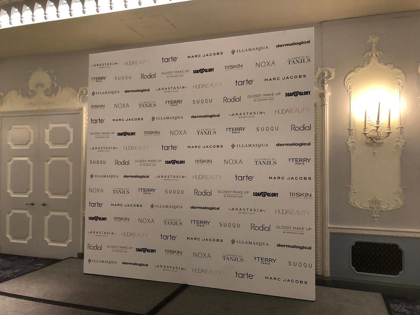 Giant white 3 by 2 metre foamboard media wall printed with rows of beauty brand logos, standing in an ornate hotel ballroom