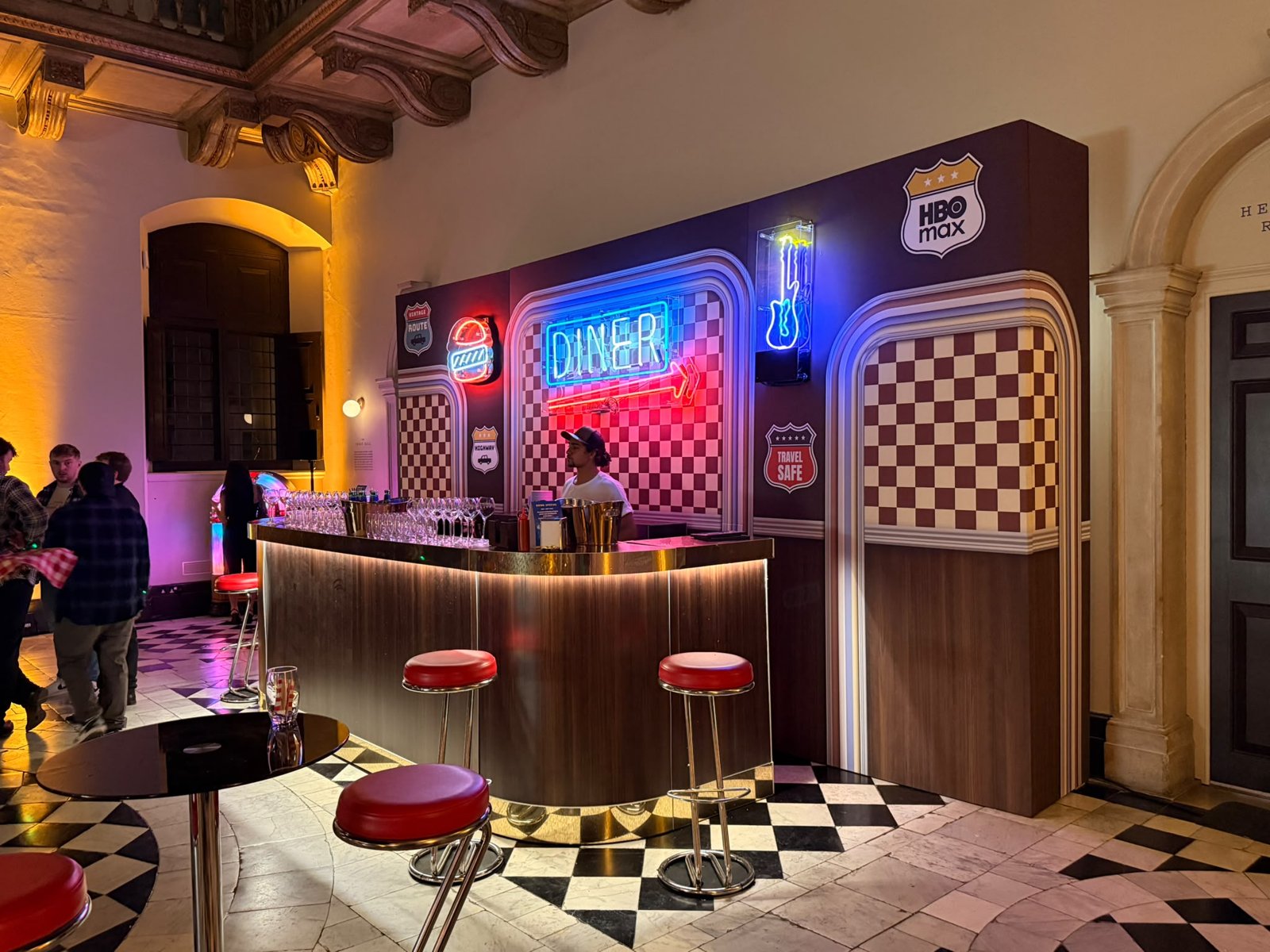 Retro American diner backdrop with chequered panels, road sign graphics and HBO Max branding behind a bar with neon signs at the Lanterns launch