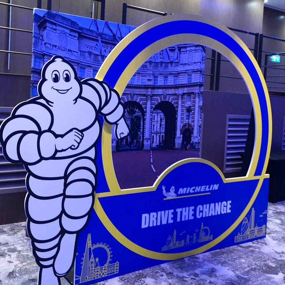 Michelin Drive the Change photo op with a cut out Michelin Man beside a blue and gold ring frame, in front of a printed Admiralty Arch backdrop