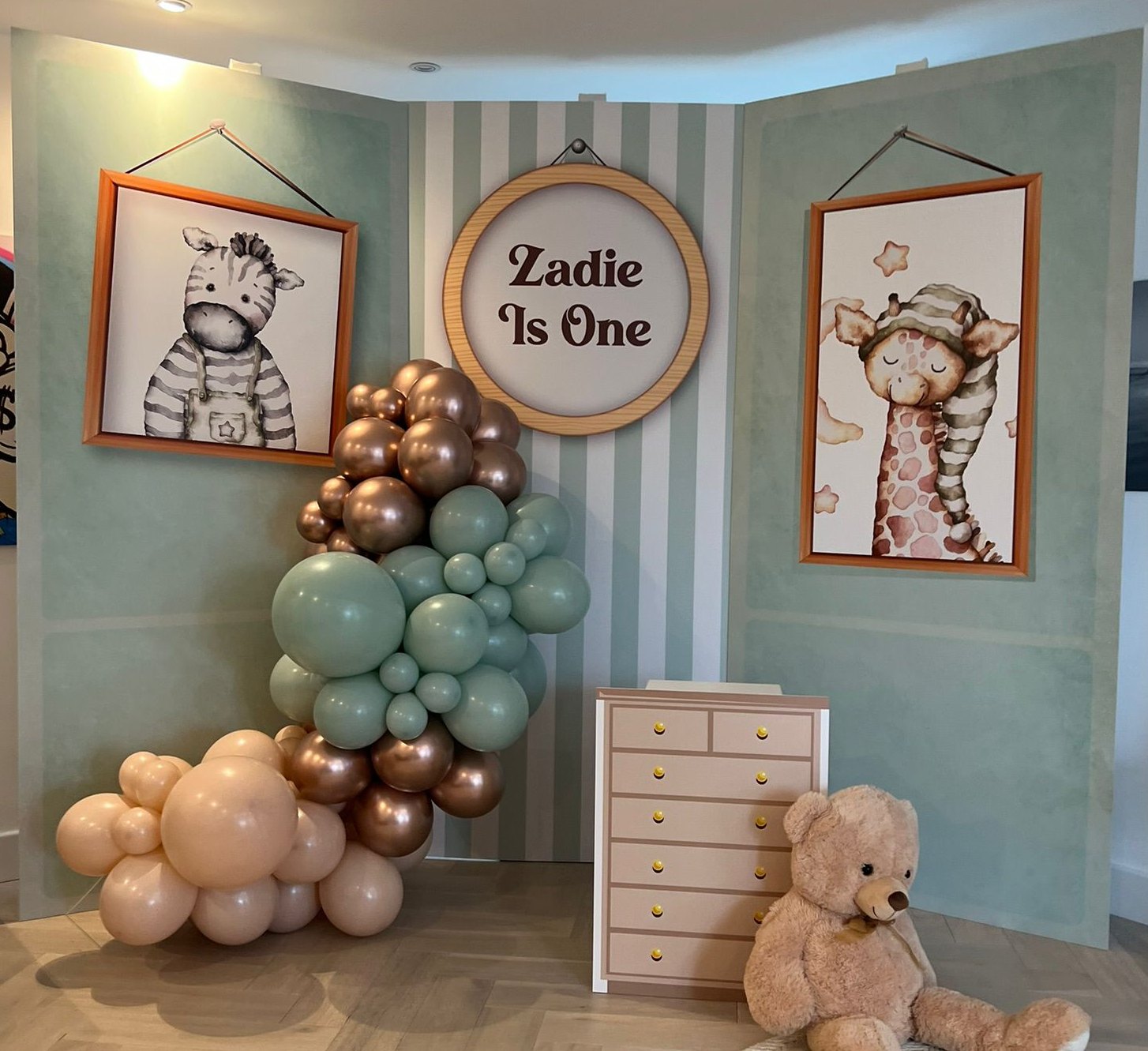 Sage green three panel nursery style first birthday backdrop with framed zebra and giraffe art, a Zadie Is One sign and a printed chest of drawers