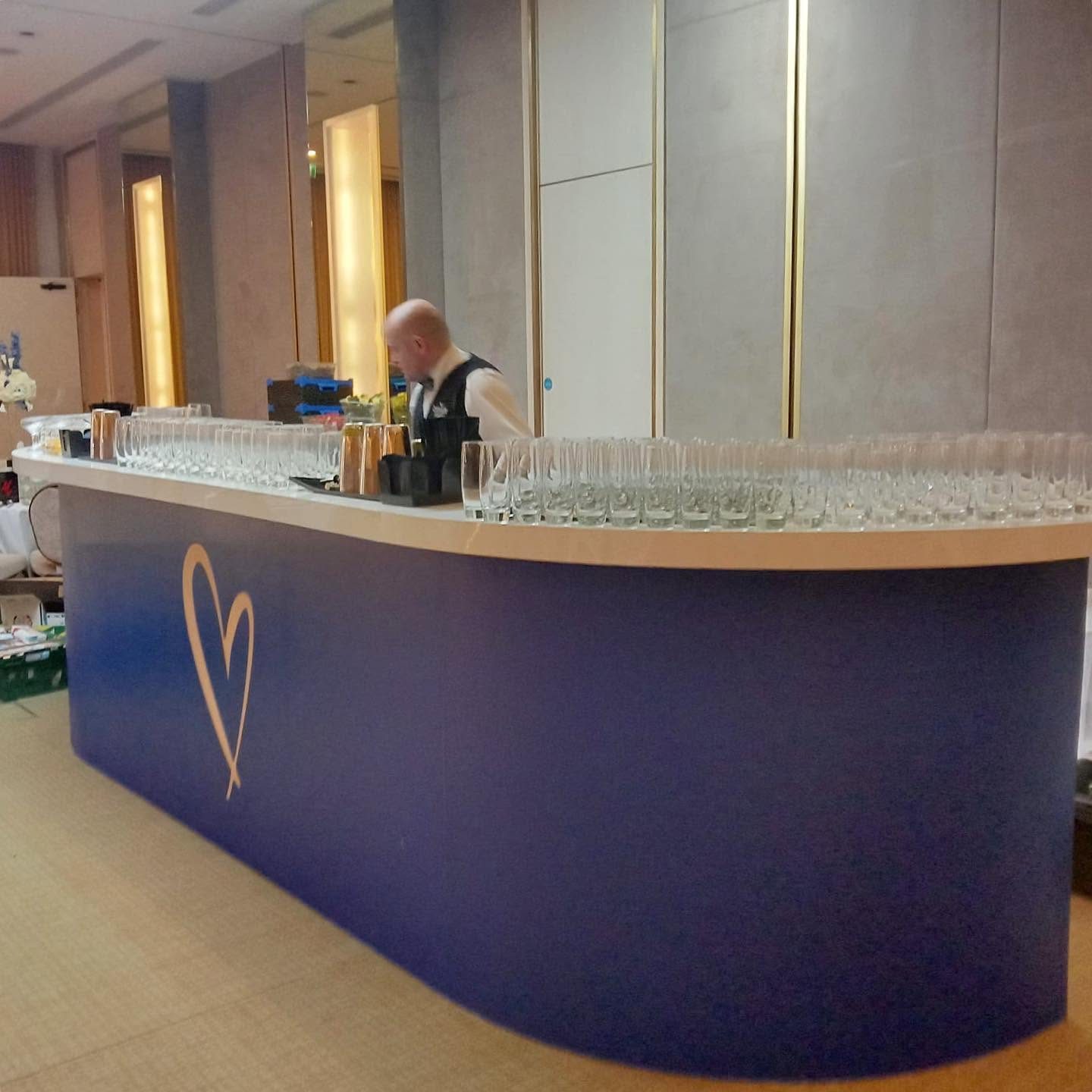Curved event bar with a deep navy printed fascia and a hand drawn gold heart, lined with rows of champagne flutes