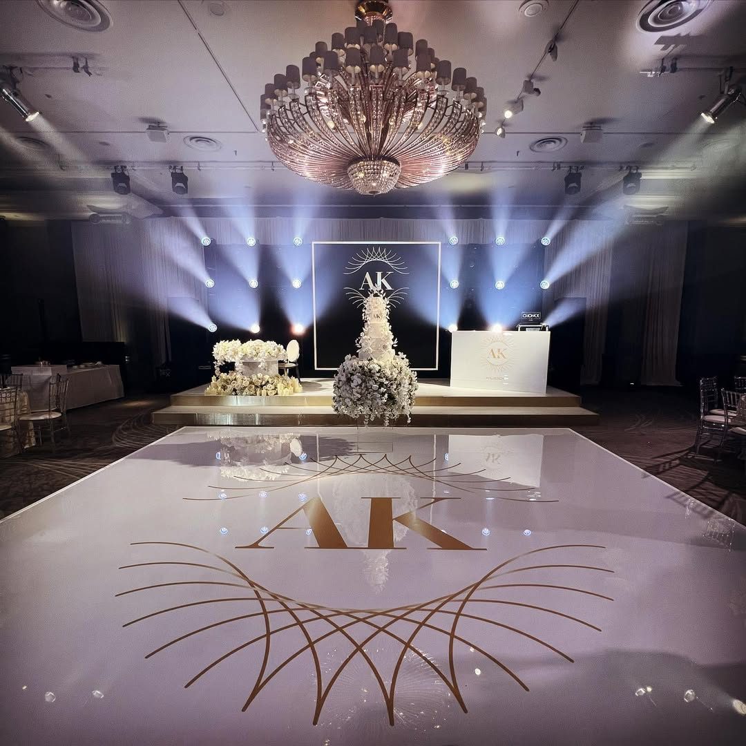 Glossy white wedding dance floor printed with a gold AK monogram and sweeping sunburst lines beneath a crystal chandelier