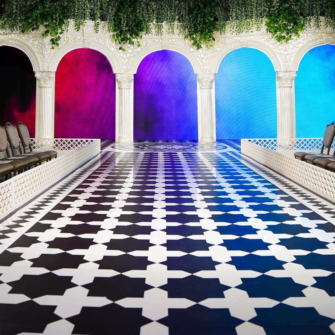 Long fashion show catwalk printed with a navy and white star and cross tile pattern, leading to white arches lit in pink, purple and blue