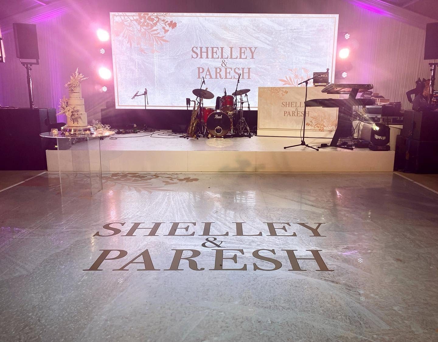White wedding dance floor printed with soft botanical artwork and Shelley and Paresh in large serif lettering in front of a band stage