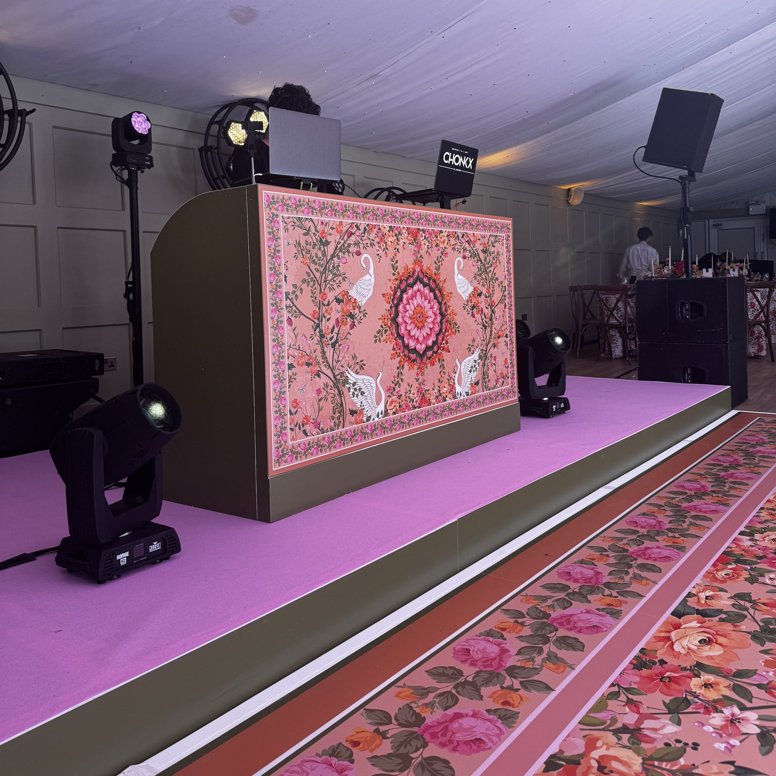 DJ booth fascia printed with a coral floral design, white cranes and a mandala centrepiece on a pink stage in a wedding marquee