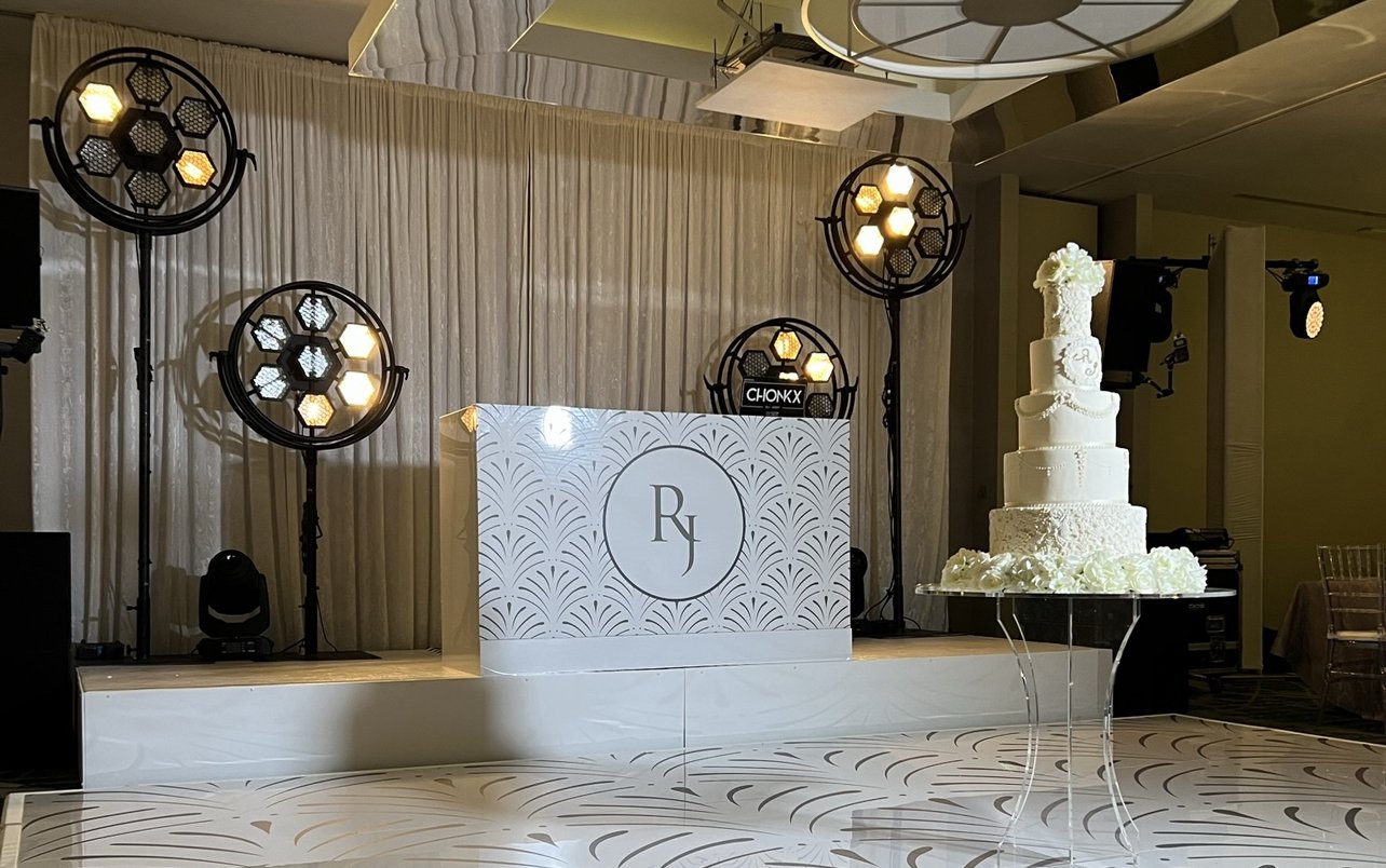 White printed DJ booth with an art deco fan pattern and RJ wedding monogram, on a stage beside a matching printed dance floor and a white tiered wedding cake