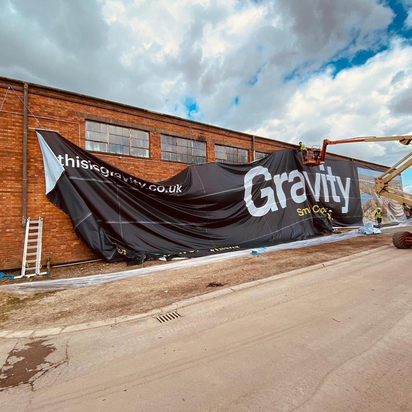 Huge black Gravity mesh banner with thisisgravity.co.uk lettering being unfurled and hoisted up the side of a brick warehouse