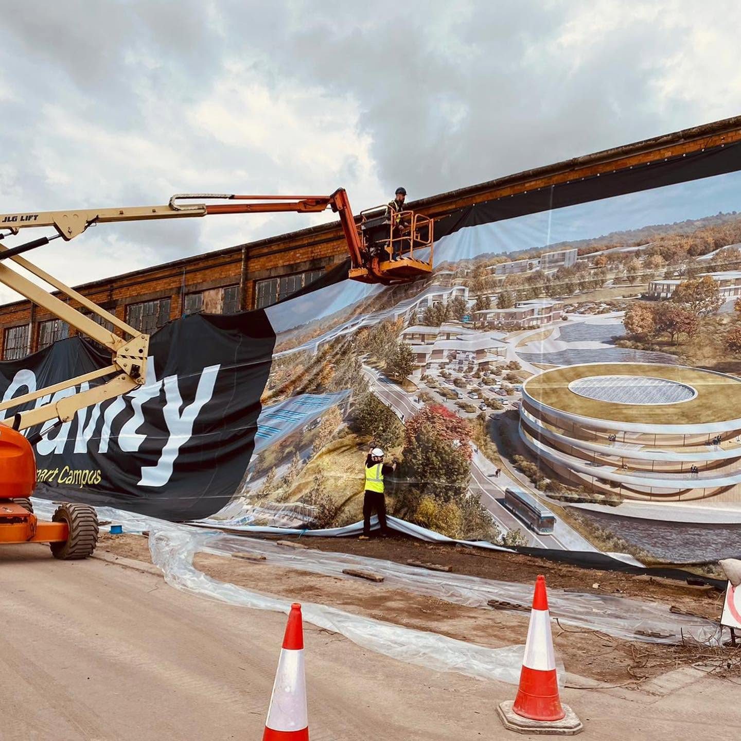 Installers on a cherry picker fitting a huge printed mesh banner showing an aerial render of the Gravity Smart Campus onto a warehouse wall