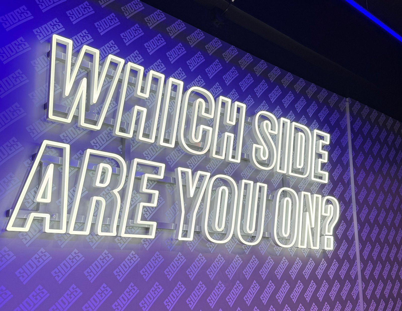 White neon sign reading Which side are you on over a purple printed vinyl wall covered in the Sides logo at the Sidemen restaurant in Hackney