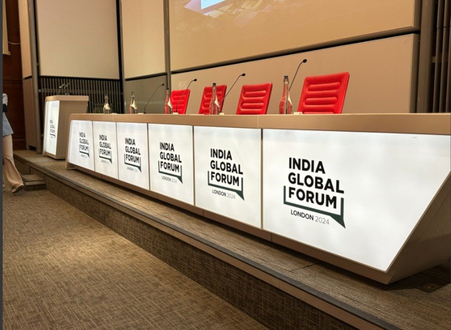 Conference panel desk and lectern wrapped in white vinyl with India Global Forum London 2024 branding beneath red speaker chairs