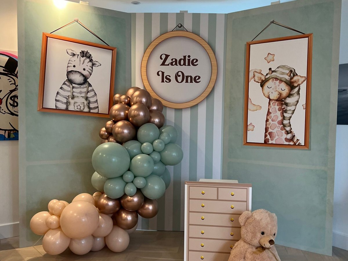 Printed first birthday backdrop with nursery animal frames, striped panel and name sign, styled with balloons