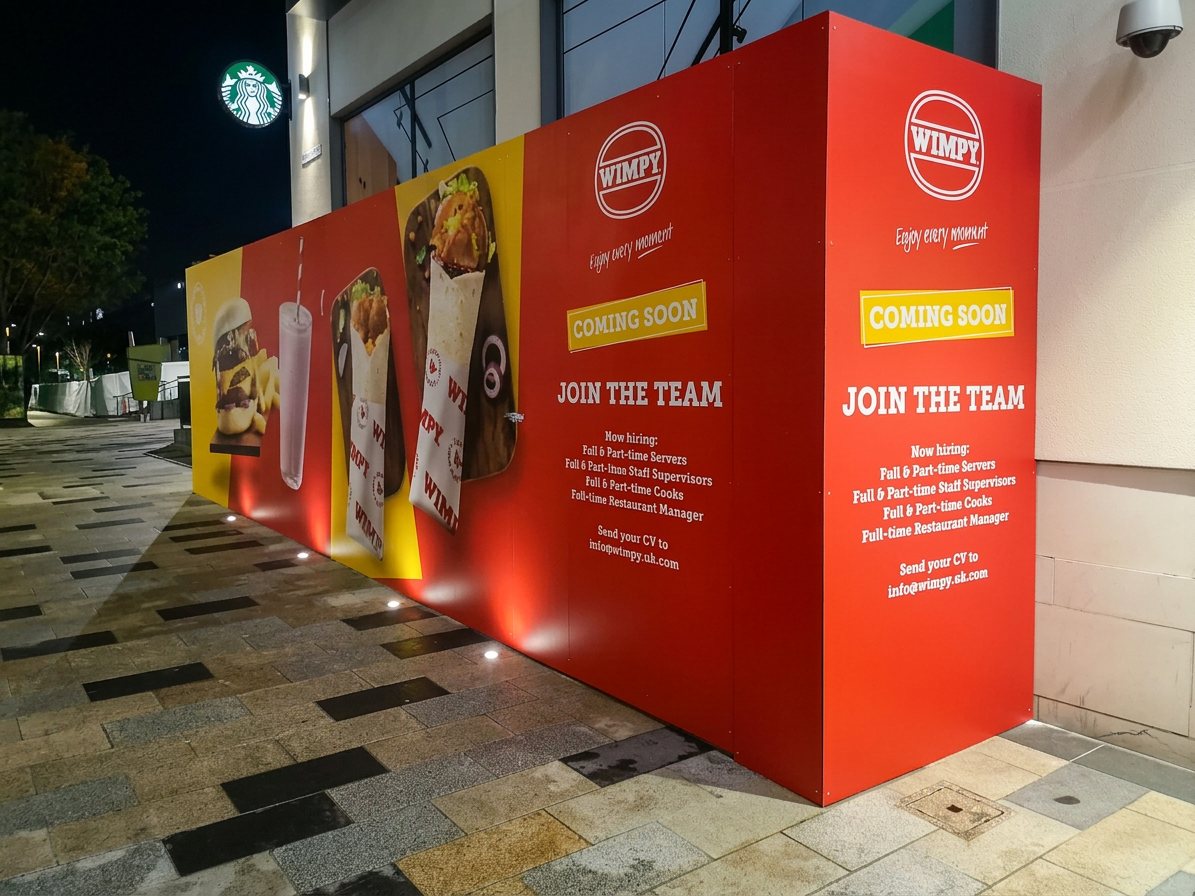 The WIMPY hoarding lit up at night with Coming Soon and Join the Team messaging